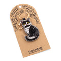 A black and white cat-shaped keychain charm with yellow eyes and a gold clasp, attached to a brown kraft paper display card labeled 'Matr Bodme'.