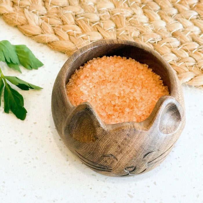 Wooden cat-shaped bowl filled with pink salt on a textured surface.
