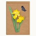 A rectangular kraft paper card featuring a handmade paper collage of two yellow daffodils with green stems and leaves, accompanied by a small blue butterfly, set against a plain white background.