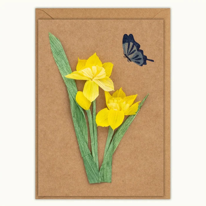 A rectangular kraft paper card featuring a handmade paper collage of two yellow daffodils with green stems and leaves, accompanied by a small blue butterfly, set against a plain white background.