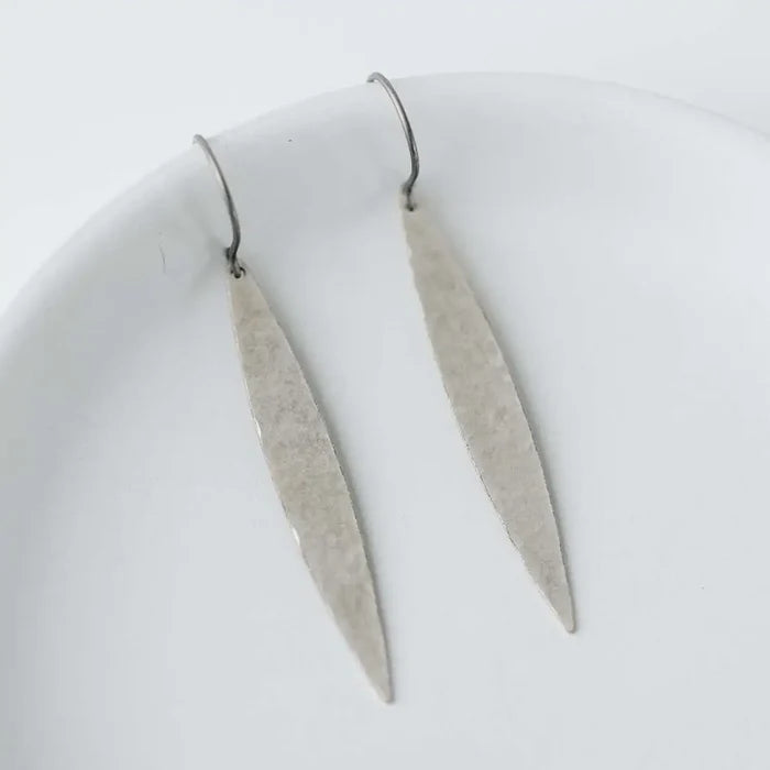 A pair of minimalist silver dangle earrings with textured, hammered elongated drop shapes and simple fishhook wires, displayed on a clean white surface.