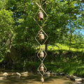 A vertical metal wind spinner with four small hanging bells, suspended from a tree branch over a shallow creek in a lush green outdoor setting.