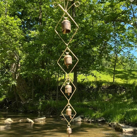 A vertical metal wind spinner with four small hanging bells, suspended from a tree branch over a shallow creek in a lush green outdoor setting.
