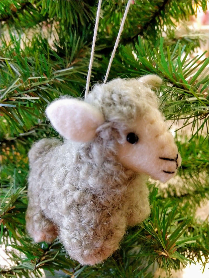 Handmade felt sheep ornament,  crafted from felted wool in beige and white colors, hanging in front of a green Christmas tree.