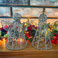 Two wire angel sculptures on a wooden surface with flowers and lights in the background.