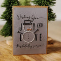 A handmade holiday greeting card with a snowman design, standing on a wooden surface with a blurred green tree in the background. The card has a gray face, a kraft brown border, and black and white embroidered details.