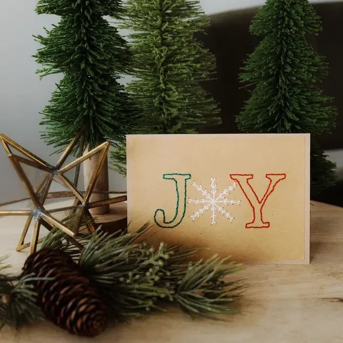 A handmade-style greeting card with the word 'JOY' embroidered in green, white, and red on a beige background, styled on a wooden surface with pine branches, a pinecone, and artificial Christmas trees.