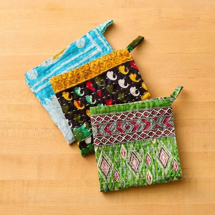 Three colorful patterned fabric dish towels or small bags arranged on a light wood surface. The front pouch is green with red, pink, and white geometric diamond patterns and a green top loop.