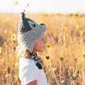 A child in profile wearing a crocheted wolf-style hat with ear-like protrusions and a braided strap, standing in a sunflower field.
