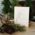 A folded greeting card with a minimalist multicolor line drawing of the Holy Family, styled on a wooden surface with pine branches, a pinecone, and artificial Christmas trees.