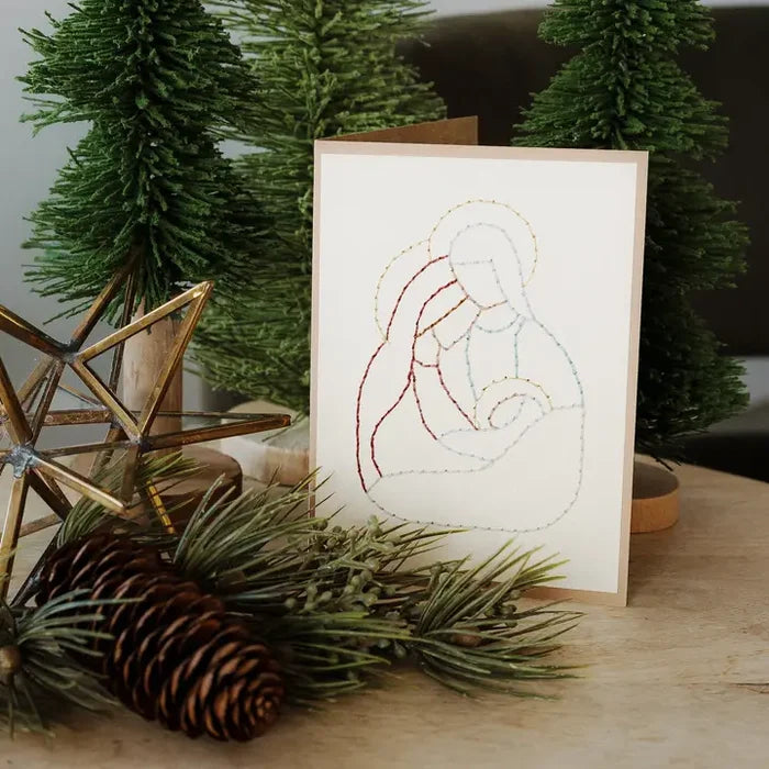 A folded greeting card with a minimalist multicolor line drawing of the Holy Family, styled on a wooden surface with pine branches, a pinecone, and artificial Christmas trees.