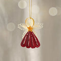 A decorative hanging ornament shaped like a stylized angel, crafted from rolled paper or quilling. The ornament has cream-colored wings, a gold ring and star-shaped center, and a deep red skirt-like lower section, suspended by a thin gold string against a soft beige background with blurred light circles.