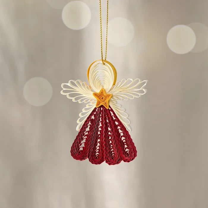 A decorative hanging ornament shaped like a stylized angel, crafted from rolled paper or quilling. The ornament has cream-colored wings, a gold ring and star-shaped center, and a deep red skirt-like lower section, suspended by a thin gold string against a soft beige background with blurred light circles.