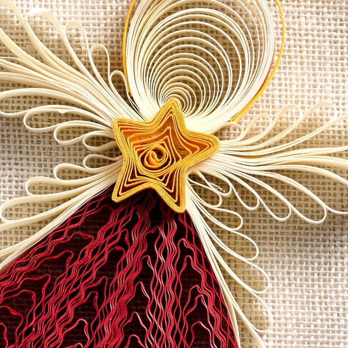 A close-up of a decorative quilled paper ornament or wall hanging on a textured beige fabric background. The design features cream-colored paper rays radiating from a gold star center, with a dark red and red patterned lower section.