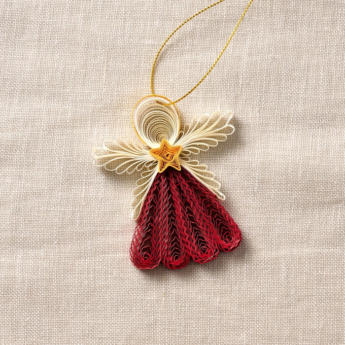 A handcrafted quilled paper angel ornament hanging from a gold string, featuring cream wings, a gold star chest, and a red dress, displayed on a neutral beige linen background.