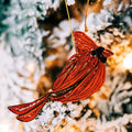 A red cardinal Christmas ornament with a gold hanging string, photographed hanging against a blurred frosted tree background with warm lights.