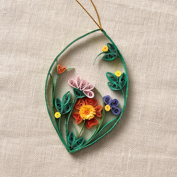 A decorative quilled paper hanging ornament shaped like a leaf with rolled floral details in green, orange, pink, purple, and yellow, displayed on a textured beige fabric background.