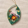 A decorative quilled paper hanging ornament shaped like a leaf with rolled floral details in green, orange, pink, purple, and yellow, displayed on a textured beige fabric background.