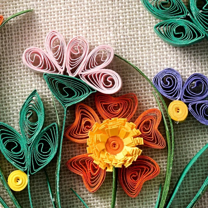 A close-up of colorful handcrafted paper flowers and leaves arranged on a textured beige fabric background. The paper flowers include pink, orange, yellow, purple, and green, with detailed layered swirls and stems.