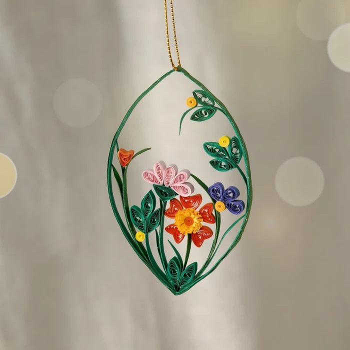 A decorative hanging ornament shaped like a teardrop, featuring a green quilled leaf outline with colorful paper flowers in pink, red, orange, purple, and yellow, suspended against a softly blurred warm gray background.