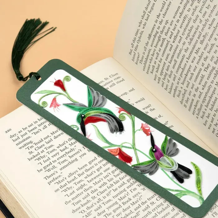 A decorative bookmark with a dark green border and tassel, featuring painted birds and floral motifs on a white background, resting on an open book.