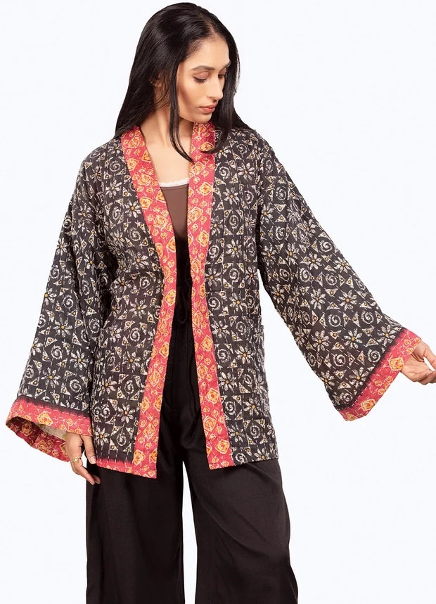 A woman modeling a black printed open-front jacket with red and gold patterned trim, paired with black pants, against a plain white background.