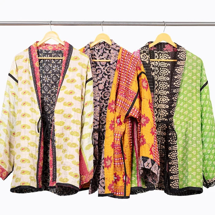 Three lightweight open-front jackets with colorful batik patterns hang on wooden hangers from a metal rack against a plain white background.
