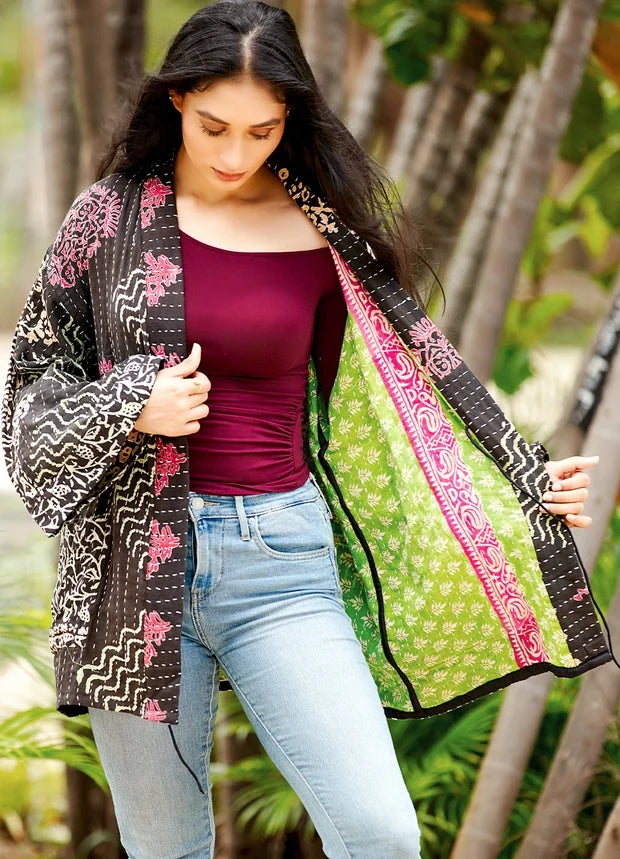A woman wearing a black and pink patterned open-front cardigan over a burgundy top and light blue jeans, standing outdoors in front of a blurred tropical background.