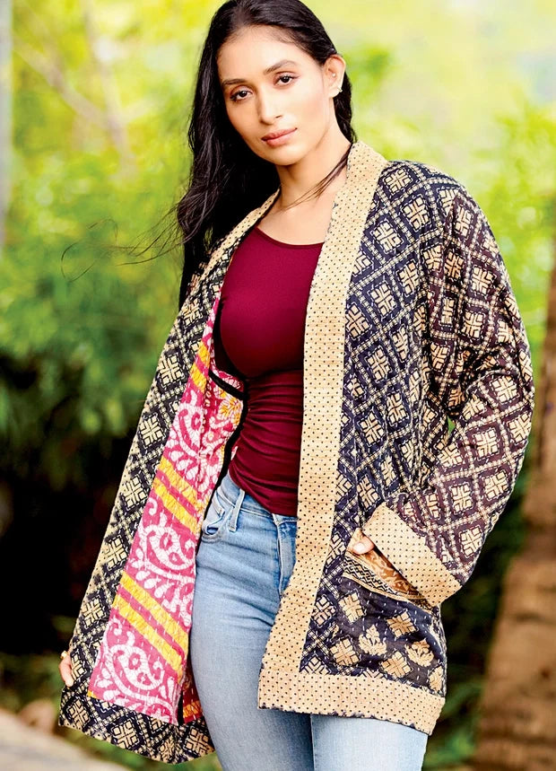 A woman wearing a patterned open-front jacket with beige and dark brown geometric designs and a pink patterned lining, styled over a burgundy top and light blue jeans against a blurred green outdoor background.