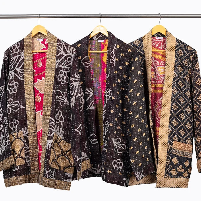 Three open lightweight quilted jackets hanging on wooden hangers from a metal rod, shown against a plain white background. Each sari jacket has a dark patterned outer fabric with beige woven trim and a bright pink inner lining with floral and geometric motifs.