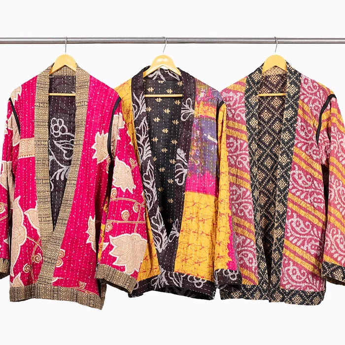 Three open-front kantha jacket hang on wooden hangers from a metal rod against a plain white background. Each cardigan features a unique patchwork design with vibrant pink, yellow, black, and beige patterns.