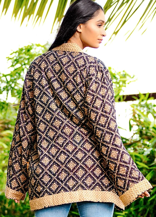 A woman wearing a loose, long-sleeve patterned cardigan or jacket with a geometric diamond design in dark brown and beige, shown from the back in an outdoor setting with green foliage.