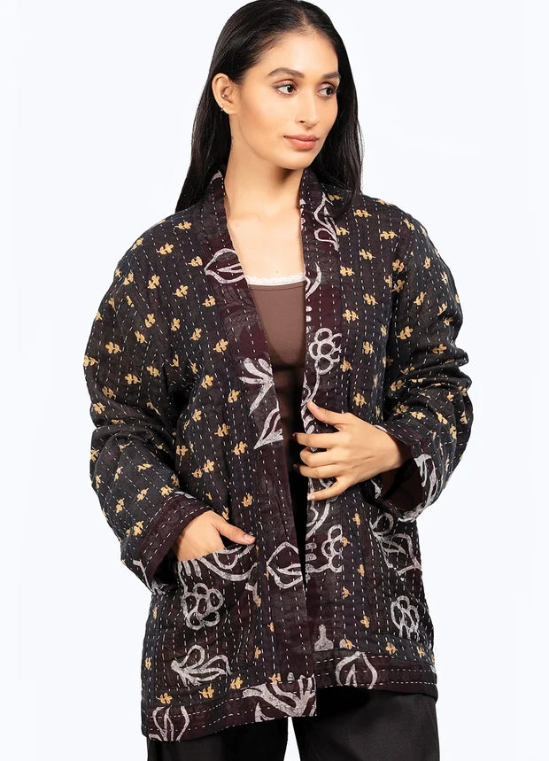 A woman modeling a black quilted jacket with white floral patterns and small gold embroidered details, worn over a brown top and black pants against a white studio background.