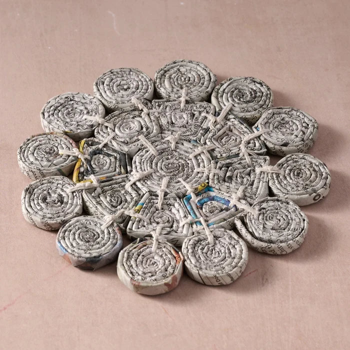 A handmade circular coiled paper trivet made from rolled newspaper strips tied together with string, displayed on a solid dusty pink background.