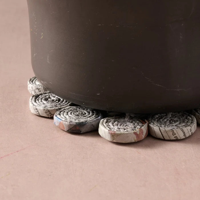 A close-up of a dark brown ceramic pot resting on several small rolled pieces of printed paper trivet on a dusty pink surface.
