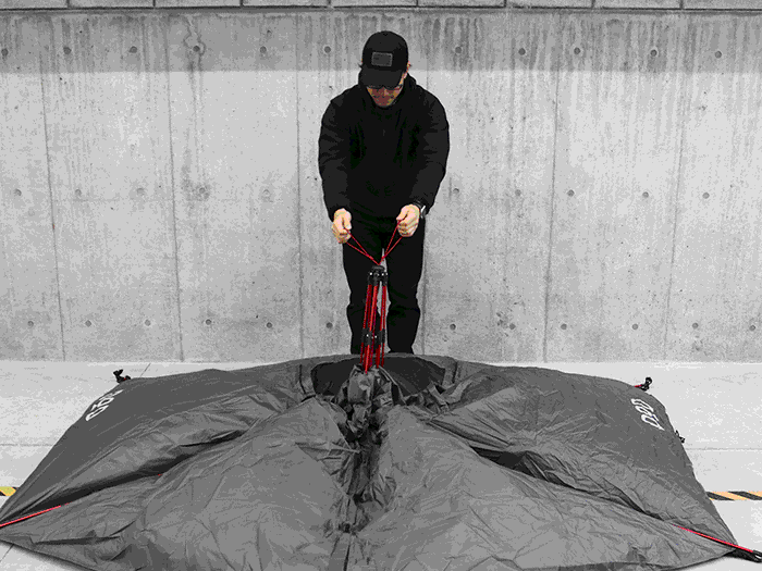 Person assembling Tomo 1Pull Dome Tent with black fabric and red poles against concrete wall