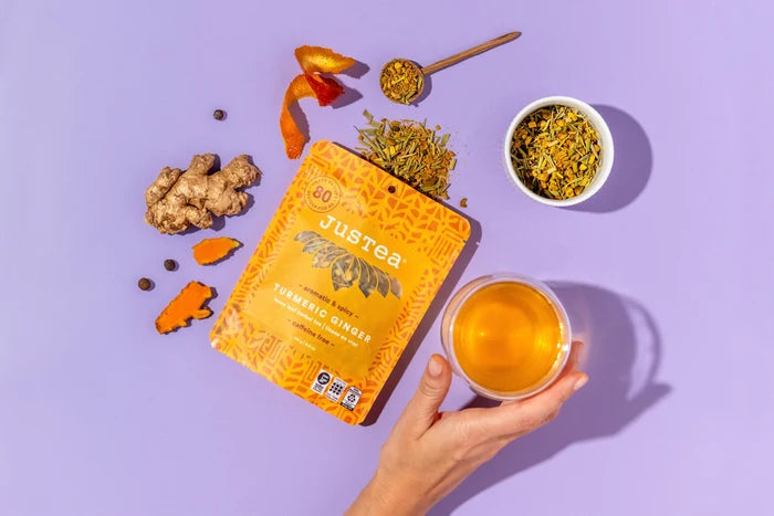 Jureea tea tumeric ginger packaging with a cup of tea, dried herbs, and a spoon on a purple background