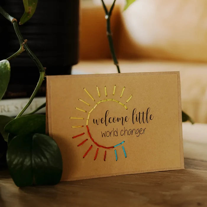 Brown greeting card with sun design and text 'welcome little world changer' on a wooden surface with plants.