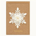 A rectangular kraft brown greeting card with a large white layered paper snowflake design in the center and gold 'Season's Greetings' text near the bottom, shown with a matching kraft envelope on a light neutral background.